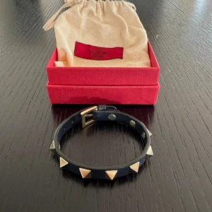 Valentino armband - Marin blått armband från Valentino i äkta skinn med ikoniska pyramidformade guldfärgade nitar runt om. Justerbart spänne i metall och levereras med originalpåse och ask. Perfekt accessoar för att ge din outfit en edgy touch.