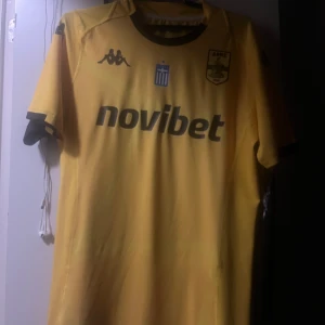 ÄKTA! Gul Aris FC matchtröja från Kappa - Gul fotbollströja från Kappa för Aris FC med svarta detaljer och tryck. Tröjan har klubbmärke, sponsorlogga och grekisk flagga på bröstet. Tillverkad i lätt och ventilerande material, perfekt för match eller träning. Klassisk passform med korta ärmar.