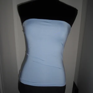 Ljusblå bandeau tubtopp - En stilren ljusblå bandeau tubtopp i stretchigt syntetmaterial. Toppen är axelbandslös och sitter tight mot kroppen, perfekt för en enkel och clean look. Passar dig som gillar minimalistisk stil och vill ha en topp som känns både modern och fräsch. Aldrig använd!