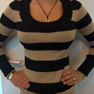 Randig stickad tröja från H&M  - Snygg stickad tröja från H&M i XS med breda svarta och guld/beiga ränder. Tröjan har en djup rundad halsringning och långa ärmar. Materialet är mjukt och stretchigt, perfekt för lager på lager under höst och vinter.