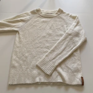 Vit stickad tröja från 157 - Mysig vit stickad tröja från 157 med rund halsringning och ribbade muddar vid ärmslut och nederkant. Tröjan har en enkel och stilren design.