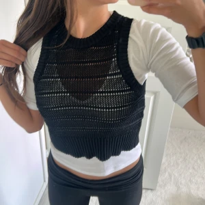 Svart stickad väst från Stylein - Säljer en svart, croppad stickad väst från Stylein i storlek S. Västen har bred ribbad kant nedtill och vid ärmhålorna, samt ett öppet, virkat mönster som ger en cool och luftig känsla. Perfekt att styla över en t-shirt eller skjorta.