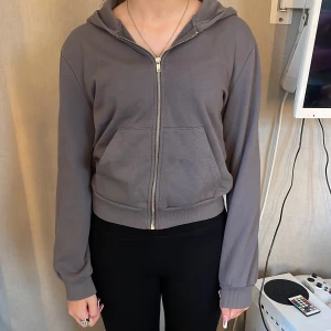 Grå hoodie med dragkedja från 157 - Mysig grå hoodie från 157 i storlek XS. Tröjan har huva, dragkedja framtill och två stora fickor. Tillverkad i mjuk bomullsmix och har ribbade muddar vid ärmslut och nederkant. Perfekt för chill dagar eller när du vill ha en enkel och stilren look.