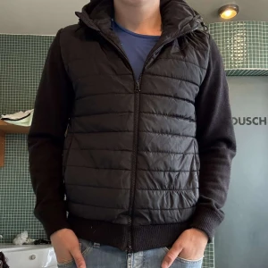 J.Lindeberg Cardigan - Svart quiltad jacka från J.Lindeberg med dragkedja framtill och ribbade muddar. Jackan har en sportig look med vadderad framsida och mjuka ärmar. Perfekt för dig som gillar stilren och enkel design. Enda deffekten är att det är hål i en av fickorna.