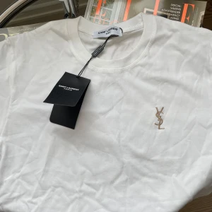 Saint Laurent T-shirt - Vit t-shirt från Saint Laurent med klassisk passform och korta ärmar. Tillverkad i mjuk bomull och prydd med broderad YSL-logga i beige på bröstet. Perfekt basic med exklusiv känsla och stilren design. Storlek S, tags medföljer. Hör av er vid frågor eller funderingar!