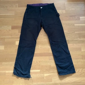 Svarta byxor Comme des Garçons Homme - Svarta byxor från Comme des Garçons Homme med raka ben och coola detaljer som mönstrat innerfoder i lila och rött. Byxorna har flera fickor och en metallknapp med logga. Tyvärr lite förstörda i hälarna som du kan se på sista bilden 