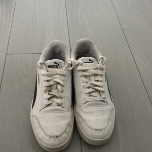 Vita Puma sneakers med svart detalj - Snygga vita Puma sneakers med svarta detaljer på sidorna och guldfärgad Puma-logga. Klassisk låg modell med snörning och perforerad tå för extra ventilation. Ovandel i syntet och textil, innersula med SoftFoam+ för skön komfort (skönast sulan jag någonsin andvändt). Perfekta för en clean och sportig stil.