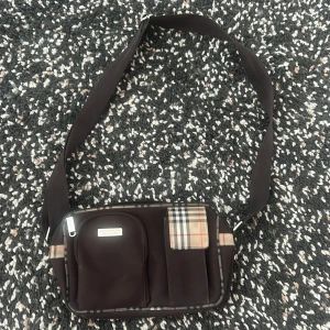 Brun axelväska från Burberry - Säljer en brun axelväska från Burberry med klassiskt rutigt mönster på detaljerna. Väskan har justerbart band, flera fack framtill och dragkedja upptill. Perfekt storlek för det viktigaste och snygg att bära crossbody.