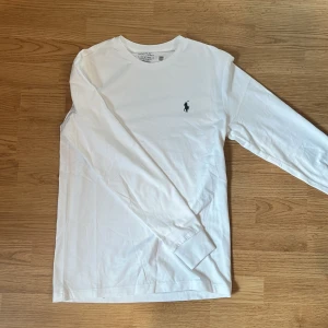 Vit långärmad tröja från Polo Ralph Lauren - Klassisk vit långärmad tröja från Polo Ralph Lauren med rund hals och ikoniska broderade loggan på bröstet. Tillverkad i mjuk bomull som känns skön mot huden. Perfekt för en clean och stilren look.