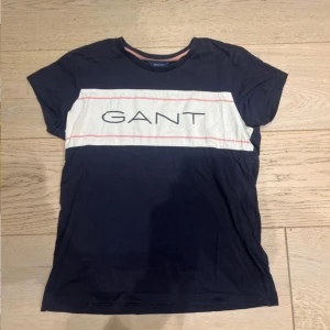 Blåvit GANT t-shirt med logga - Snygg t-shirt från GANT i marinblått med vit panel och rosa linjer över bröstet. Stor GANT-logga framtill. Klassisk passform och rund halsringning med rosa detalj. Perfekt för en chill och stilren look.