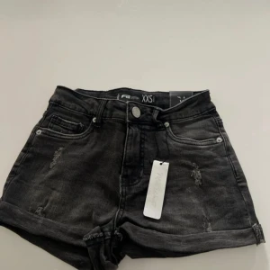 Svarta jeansshorts från FB Sister - Snygga svarta jeansshorts från FB Sister i storlek XXS. Modellen är högmidjad med upprullade benslut och slitna detaljer framtill för en cool look. Klassisk femficksdesign och knappgylf. Perfekta för sommaren när du vill ha en edgy stil.