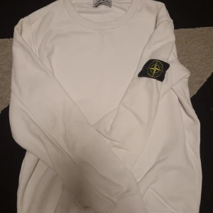 Vit sweatshirt från Stone Island - Vit sweatshirt från Stone Island med klassisk rund hals och ikonisk logotyp-patch på vänster ärm. Tröjan är i mjuk bomull och har en clean, stilren look som passar perfekt till streetwear. Långärmad modell med relaxed fit.