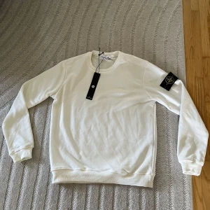 Vit sweatshirt från Stone Island M - Säljer en clean vit sweatshirt från Stone Island i storlek M. Tröjan har rund hals, långa ärmar och den klassiska loggan på vänster ärm. Tillverkad i mjuk bomull för skön känsla och stilren look. Perfekt för dig som gillar minimalistiskt mode.