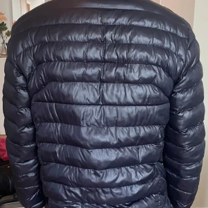 Svart dunjacka Polo Ralph Lauren - Snygg svart pufferjacka från Polo Ralph Lauren med klassisk quiltad design och dragkedja framtill. Jackan har hög krage, två sidofickor med dragkedja och diskret logga på bröstet. Perfekt för kyliga dagar och enkel att matcha med allt.