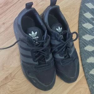 Svarta Adidas ZX sneakers strl 40 - Snygga svarta Adidas ZX sneakers med klassiska tre ränder på sidan. Ovandel i mesh och syntet med detaljer i mocka. Skorna har snörning och platt sula, perfekt för en sportig och stilren look.