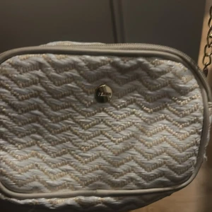 Beige och vit axelväska med kedja - Snygg axelväska från Lindex i beige och vitt med vågigt mönster. Väskan har en guldfärgad kedjerem och rundad form. Materialet är flätat och ger en somrig känsla. Perfekt för dig som vill ha en stilren och trendig accessoar.