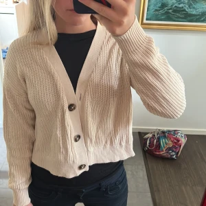 Beige kofta från Selected Femme - Snygg beige kofta från Selected Femme i stickad bomull. Modellen är croppad med v-ringning och stora knappar framtill. Perfekt att slänga över en t-shirt eller linne. Långärmad och med ribbade muddar för en chill vibe.