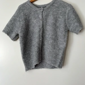 Grå boucle kofta från Gina Tricot - Supermjuk och fluffig gråmelerad kofta från Gina Tricot i bouclé-stickat material. Kortärmad modell med rund hals och silvriga knappar framtill. Perfekt för lager-på-lager och ger en chill men ändå stilren vibe.