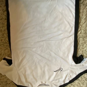 Vit och svart Nike fotbollströja - Säljer en klassisk vit fotbollströja från Nike med svarta detaljer längs sidorna och axlarna. Tröjan har korta ärmar, rund hals och är tillverkad i ett lätt och ventilerande funktionsmaterial som passar perfekt för träning eller match.