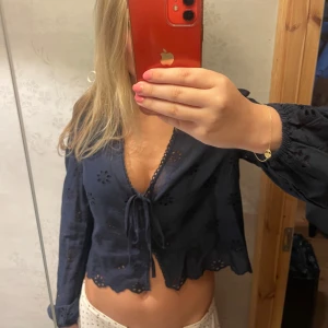 Mörkblå blus med knyt och broderi - Säljer en mörkblå blus med v-ringning och knyt framtill. Blusen har långa ärmar med lätt puff och fina broderade blommor som ger en cool och romantisk vibe. Perfekt till jeans eller kjol för en trendig look. Prislappen är kvar!!
