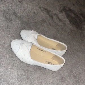 Vita ballerinaskor med knut från Clou - Söta vita ballerinaskor från Clou i storlek 37. Skorna har en twistad knut framtill och är täckta av små prickar i tyg för en extra detalj. Perfekta för dig som gillar en clean och feminin stil.
