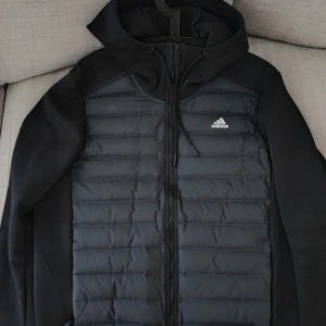 Svart hybrid pufferjacka från Adidas med huva - Svart Adidas Varilite Hybrid pufferjacka med huva och dragkedja framtill. Jackan har quiltad framsida och släta ärmar, samt Adidas-logga på bröstet. Perfekt för kyliga dagar och har en sportig vibe, också på spela golf. Långärmad och med normal passform.