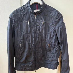 Mörkblå vindjacka från Moncler - Snygg mörkblå vindjacka från Moncler med hög krage och flera dragkedjeförsedda fickor framtill. Jackan har en stilren design med quiltade detaljer på axlar och ärmar. Perfekt för dig som vill ha en lätt och sportig jacka med exklusiv känsla.Modellen är kort och modellen på jackan heter DONATTIEN GIUBBOTTO. Hör gärna av dig om du har några frågor 😁