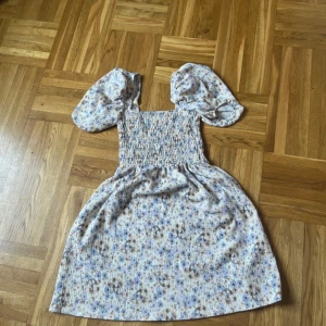 Blommig kortklänning med puffärm - Supersöt kortklänning med blått och beige blommönster, smockad överdel och stora puffärmar. Perfekt för varma dagar och ger en romantisk vibe. Lätt och luftig känsla, passar dig som gillar en feminin stil.