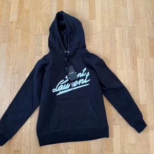 Saint Laurent hoodie  - Svart hoodie från Saint Laurent med vit logotyp framtill i coolt tryck. Klassisk känguruficka, snörning i huvan och långa ärmar. Perfekt för dig som gillar streetstyle och vill ha något trendigt och bekvämt. (iykyk)
