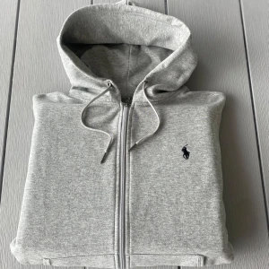 Grå hoodie – Polo Ralph Lauren – Strl S - Klassisk ljusgrå hoodie från Polo Ralph Lauren i storlek S (170/182A). 🧵 Broderad logga i mörkblått 🧥 Dragkedja framtill 🤍 Mjuk och bekväm – perfekt till vardags 📍 Fint skick – knappt använd  Perfekt basic att ha året runt!