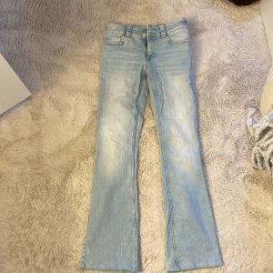 Ljusblå bootcut jeans med fickdetaljer - Säljer ett par ljusblå bootcut jeans med snygg tvätt och coola bakfickor med knappdetaljer. Jeansen är lågmidjade, har klassisk femficksdesign och dragkedja med dubbla knappar. Perfekta för en avslappnad och trendig look.