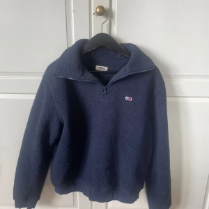 Mörkblå fleece-tröja Tommy Jeans - Mysig mörkblå fleece-tröja från Tommy Jeans med halv dragkedja och broderad logga på bröstet. Tröjan har hög krage och långa ärmar, perfekt för kyliga dagar. Enkel och stilren design som funkar till det mesta.