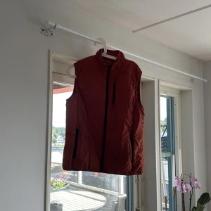Rostbrun quiltad dunväst - Säljer en rostbrun quiltad dunväst med hög krage och svarta dragkedjor. Västen har tre fickor med dragkedja framtill och en stilren, sportig look. Perfekt att slänga över en hoodie eller tröja när det är kyligt ute.