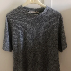 Grå ribbad stickad tröja - Säljer en grå ribbad stickad tröja med korta ärmar och rund hals. Tröjan har en enkel och stilren look, perfekt för lager-på-lager eller att bära som den är. Mjuk och skön känsla, passar till många olika outfits.