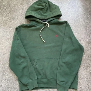 Grön hoodie från Polo Ralph Lauren - Mörkgrön hoodie från Polo Ralph Lauren med klassisk huva och snörning. Tröjan har en broderad rosa logga på bröstet, känguruficka och är tillverkad i mjuk bomullsmix. Perfekt för chill dagar eller när du vill ha en clean streetstyle.