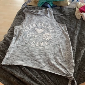 Gråmelerad linne-topp H&M med tryck - Säljer en gråmelerad ärmlös linne-topp från H&M med vitt tryck 'TEAM SELFIE CAMP' och emojis framtill. Toppen har knyt-detalj i ena sidan och är perfekt för sommarens chill dagar. Supermjuk och lätt att matcha med jeans eller shorts.