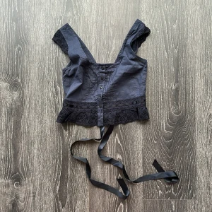 Marinblå korsettopp med spets och snörning - Supersnygg marinblå korsettopp med broderade spetsdetaljer och knappar framtill. Toppen har breda axelband, volangkant nertill och dekorativ snörning i satinband på ryggen. Perfekt för dig som vill ha en edgy och romantisk vibe.