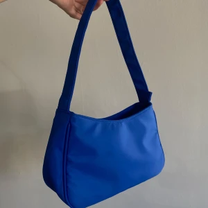 Blå axelväska i syntetmaterial - Snygg blå axelväska med enkel och stilren design. Väskan har en mjuk, rundad form och är tillverkad i syntetmaterial. Stängs med dragkedja upptill och har ett bredare axelband som gör den bekväm att bära. P