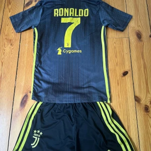Juventus Ronaldo matchtröja Adidas - Juventus fotbollströja med Ronaldo 7-tryck, svart med gula detaljer och diskret prickigt mönster. Officiella loggor från Adidas, Jeep och Juventus. Kortärmad modell i lätt och ventilerande material, perfekt för fotbollsplanen eller att bära som supporter.