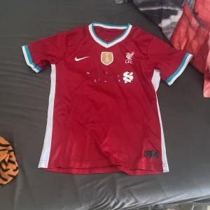 Liverpool FC röd Nike matchtröja - Liverpool FC matchtröja i rött med vita och turkosa detaljer på ärmslut och krage. Tröjan har klubbmärke och Nike-logga på bröstet samt FIFA-märke. Tillverkad i lätt och ventilerande material, perfekt för fotboll eller supporterliv.