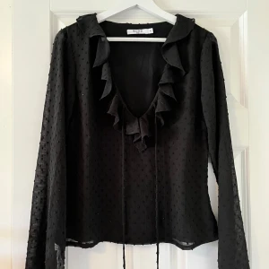 Svart blus med volang från NA-KD - Svart blus från NA-KD med lång ärm och v-ringning. Blusen har volangdetaljer längs ringningen och knytband framtill. Tunt, lätt material med diskret prickig struktur som ger en cool textur. Perfekt för dig som gillar trendiga detaljer och en feminin touch.