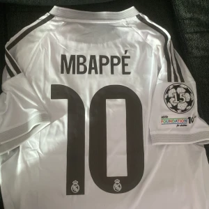 Real Madrid Mbappé 10 fotbollströja - Säljer en Real Madrid fotbollströja med Mbappé och nummer 10 på ryggen. Tröjan är vit med svarta detaljer, gula inslag och broderat klubbmärke. Champions League-märke på ärmen och Aeroready-material för bästa komfort. Perfekt för dig som älskar fotboll och Real! Storlek M