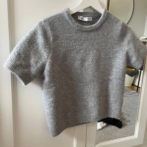 Grå stickad kortärmad tröja Zara - Supermjuk grå stickad tröja från Zara med korta ärmar och rund hals. Perfekt för lager-på-lager och har en clean, enkel look med ribbade kanter vid ärmslut och nederkant. Passar dig som gillar minimalistisk stil.