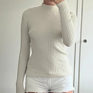 Ribbad vit långärmad topp med öppen rygg - Supersnygg ribbad vit långärmad topp med hög hals och öppen rygg som knyts med band i nacken. Figurnära passform och stretchigt material, perfekt för att lyfta din outfit med en unik detalj i ryggen.