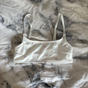 Vit blommig bikiniöverdel lager 157 - Supersöt bikiniöverdel från lager 157 i modellen Winnie Top. Den är vit med små blommiga detaljer i rosa och grönt, har smala justerbara axelband och en enkel, sportig form. Perfekt för lata dagar vid stranden eller poolen.