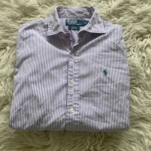 Lila randig skjorta från Polo Ralph Lauren - Säljer en lila och vit randig skjorta från Polo Ralph Lauren, modell Stanton Custom Fit. Skjortan har klassisk krage, långa ärmar och knäppning framtill. Ikoniska gröna loggan broderad på bröstet. Perfekt för dig som gillar stilrena och fräscha plagg.