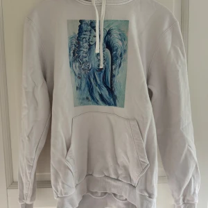 Vit hoodie  - Vit hoodie med stor ficka framtill och snörning i huvan. På bröstet finns ett blått konstnärligt tryck av en kvinna med lockigt hår. Hoodien har långa ärmar och ribbade muddar. Perfekt för dig som gillar unika prints och chill stil.