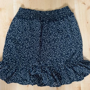 Mörkblå kortkjol med volang - Supersöt mörkblå kortkjol med vitt mönster och bred resår i midjan. Kjolen har volang längst ner som ger en extra touch och flowig känsla.