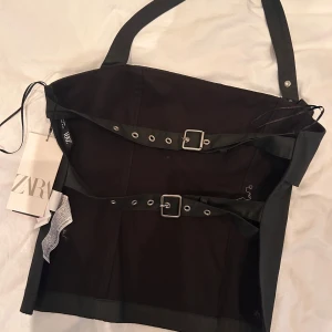 Svart kort klänning med bältesdetaljer Zara - Supersnygg svart kort klänning från Zara med coola bältesdetaljer och spännen framtill. Axelbandsmodell med raka linjer och minimalistisk look. Perfekt för dig som gillar edgy stil och vill sticka ut. Materialet känns mjukt och följsamt.