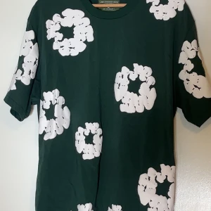 Grön t-shirt med blommigt print Denim Tears - Snygg grön t-shirt från Denim Tears med stora vita blommiga mönster över hela plagget. Klassisk passform med korta ärmar och rund hals. Tillverkad i mjuk bomull och perfekt för dig som vill sticka ut med en unik stil.